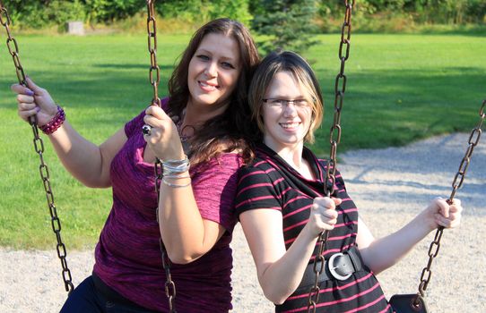 Attractive mother and daughter family sitting smiling outdoors sitting on a swing in the playground