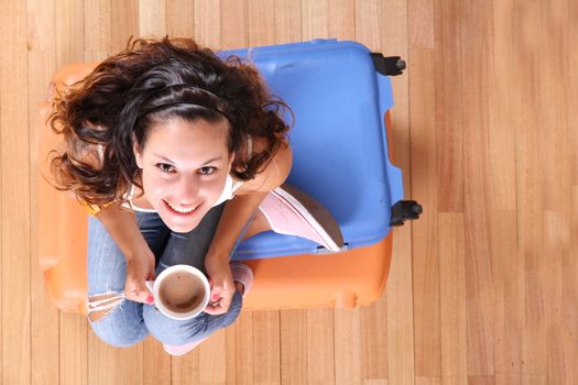 A young woman sitting on a stack of suitcases while drinking coffee and waiting for the departure to vacations.