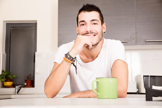 A young man sitting in the kitchen and drinking coffee.