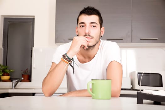 A young man sitting in the kitchen and drinking coffee.