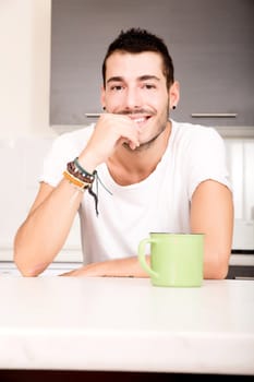 A young man sitting in the kitchen and drinking coffee.