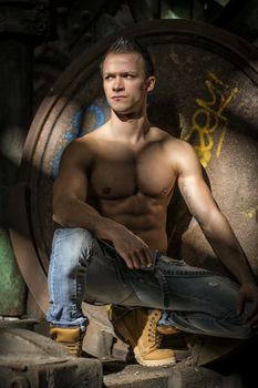 Muscular shirtless young man in abandoned industrial warehouse kneeling, looking away