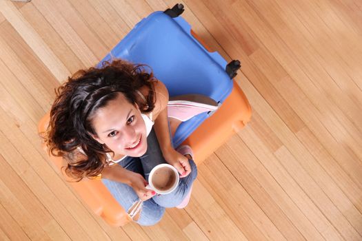 A young woman sitting on a stack of suitcases while drinking coffee and waiting for the departure to vacations.