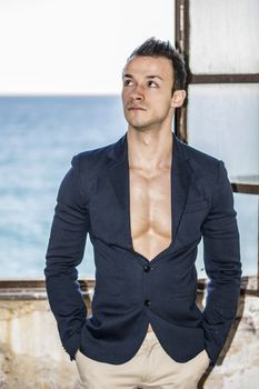 Muscular young man with jacket on naked torso, window on sea behind him
