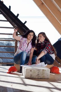 Two young women workers sitting on under construction roof, and control building in accordance with drawing