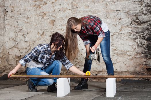 Two young long-haired woman cut iron pipe with angle grinder during the repair an old house