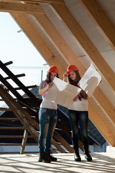 Two young women workers standing on under construction roof, and control building in accordance with drawing