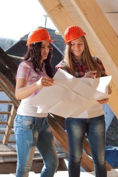 Two young women workers standing on under construction roof, and control building in accordance with drawing