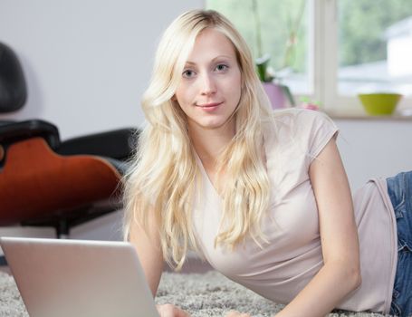 Relaxed woman is using the laptop in her living room, lying on the carpet