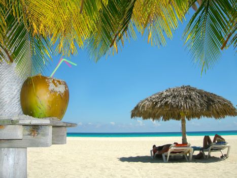 People relaxing under tropical huts with coconut and palm leaves in the foreground in a Cuban beach