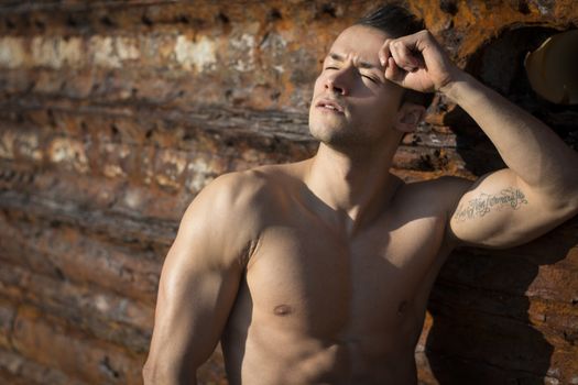Muscular shirtless young man sunbathing on rock by rusty metal wall, eyes closed