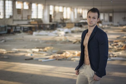 Muscular young man with jacket on naked torso in empty abandoned workplace