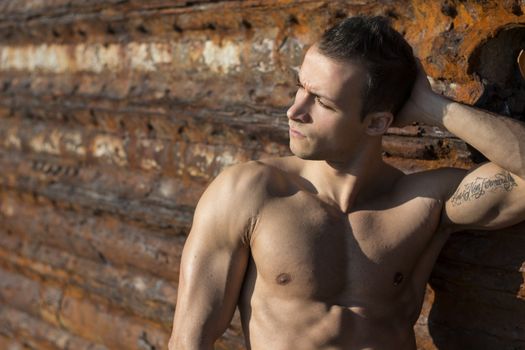 Muscular shirtless young man sunbathing on rock by rusty metal wall, looking away