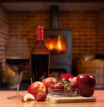 Red wine in bottle and glass with apple and strawberries on wooden table in front of roaring fire inside wood burning stove in brick fireplace