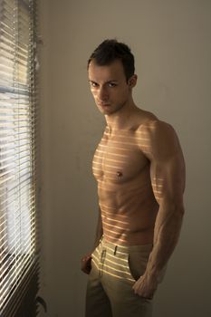 Muscular shirtless man lit by sun behind venetian blinds looking at camera