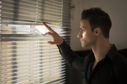 Profile of young man peeking through venetian blinds in a dark room