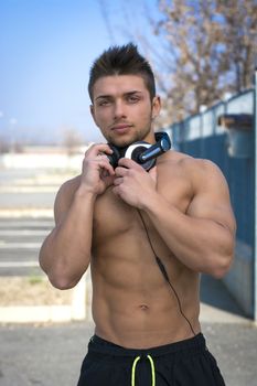 Muscular shirtless young man outdoors with headphones smiling and looking at camera