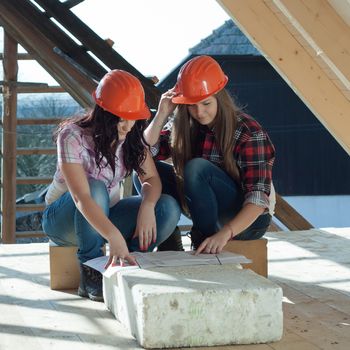 Two young women workers sitting on under construction roof, and control building in accordance with drawing