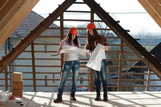 Two young women workers standing on under construction roof, and control building in accordance with drawing