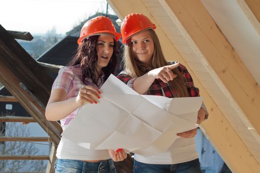 Two young women workers standing on under construction roof, and control building in accordance with drawing