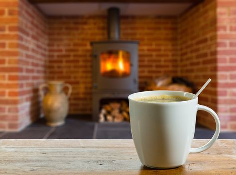 Coffee cup on wooden table in front of roaring fire inside wood burning stove in brick fireplace