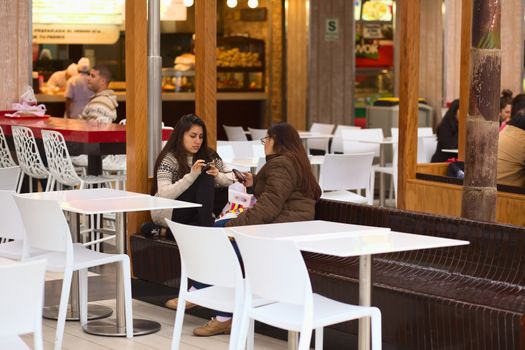 LIMA, PERU - JULY 23, 2013: Unidentified young women sitting with cell phones on bench in the food court of the shopping mall Larcomar on July 23, 2013 in Miraflores, Lima, Peru. Larcomar is a very popular shopping mall in Lima built into the steep coast of Miraflores overlooking the Pacific Ocean. 