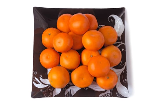 Large ripe oranges are located on a dish made of dark glass with ornament. Presented on a white background