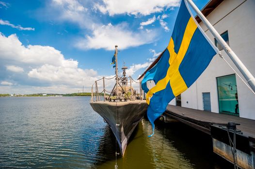 Blue and yellow flag of Sweden blowing in breese with warship moored in harobour, in background visible blue sky and clouds.
