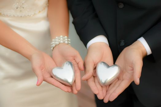 Man and woman, elegantly dressed, wearing wedding clothes, holding symbolic silver hearts in their hands, softfocus, visible unrecognizable fragments of body, model release not required.
