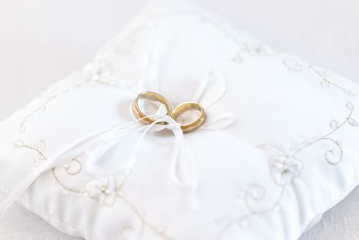 Two wedding rings on embroided white cushion, tied by white ribbon.