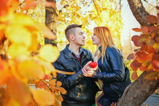 Young couple holding a red heart standing between trees autumn park