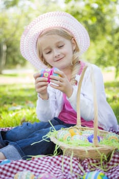 Cute Young Girl on Picnic Blanket Wearing Hat Enjoys Her Easter Eggs Outside in the Park.