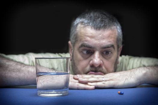 Frustrated young man sitting at the table and looking at the pills laying on it 