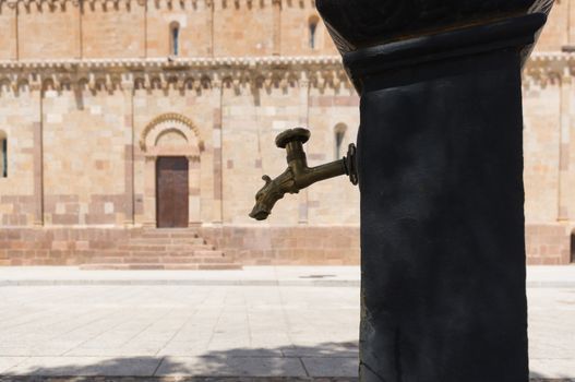 Drinking fountain with faucet-shaped dragon. In the background, a Romanesque church. (Sardinia)