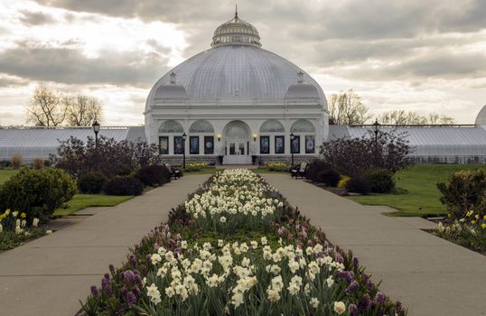 Rows of well maintained flower beds line the sidewalk leading up to an old domed and elaborate botanical garden structure. 