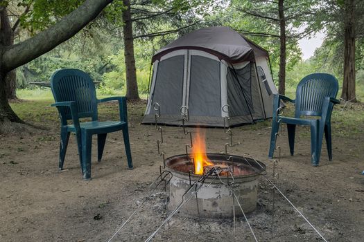 Pitched tent, crackling campfire, two chairs, and metal roasting sticks. All under a canopy of trees.