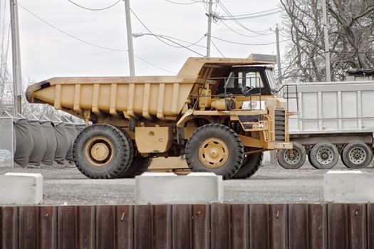 A big-wheeled construction truck sits parked among other vehicles and machinery at a factory lot along the canal. The background color is absent so the giant yellow truck stands out. 