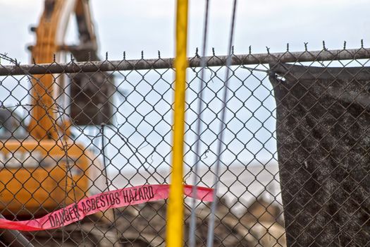 A red hazard banner hangs loosely on a chain link fence. Focus is on the fence and warning. Working machinery is barely seen in the background at this dangerous factory demolition site. 