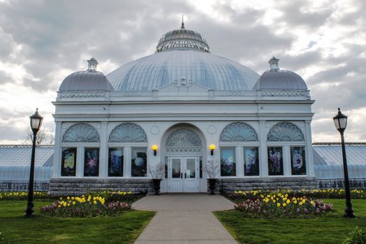 Magnificent greenhouse structure with tri domed roof and beautiful window designs.  An assortment of flowers borders the conservatory entrance walk. Silver clouds float overhead. 