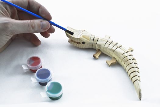 Hand holds a paint brush near a wooden toy crocodile. A set is paints are nearby.