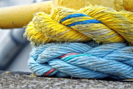 Two thick boating ropes wrapped around structure. Focus is on the fraying yellow and blue ropes with a shallow depth of field.