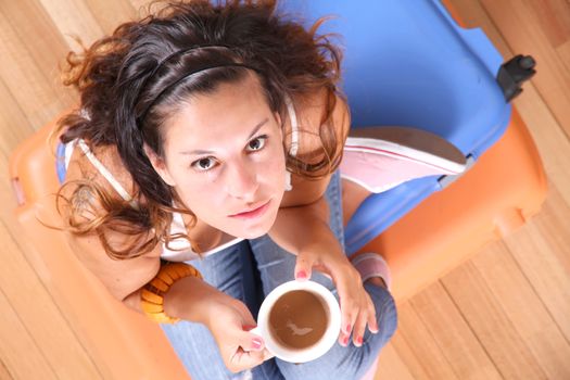 A young woman sitting on a stack of suitcases while drinking coffee and waiting for the departure to vacations.