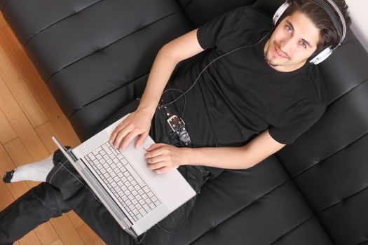 A young hispanic man with a laptop on the Sofa listening Music with headphones.