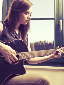 Photo of a woman playing an acoustic guitar by a window. Heavily filtered.