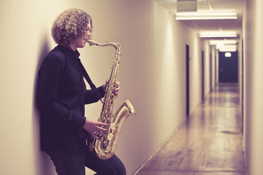 Photo of a young man playing the saxophone in a hallway. Heavily filtered.
