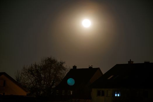 The moon being reflected by the camera lense and now visible in a second instance over buildings and trees