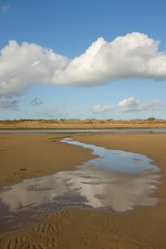A clean sandy beach with water flowing to a river, the sky is refected in the water, a large cloud cuts across a clear blue sky.