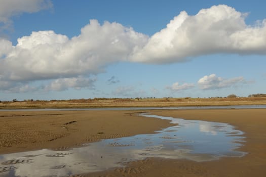 A clean sandy beach with water flowing to a river, the sky is refected in the water, a large cloud cuts across a clear blue sky.