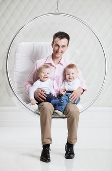 Young Caucasian man with two babies having fun while sitting in swinging hanging chair
