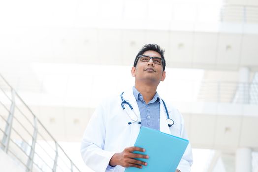Portrait of an Asian Indian male medical doctor in lab uniform holding file folder standing in front of hospital.